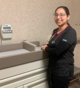 Tiffany Velasco, CCMA, stands next to pediatric weight station in black scrubs.